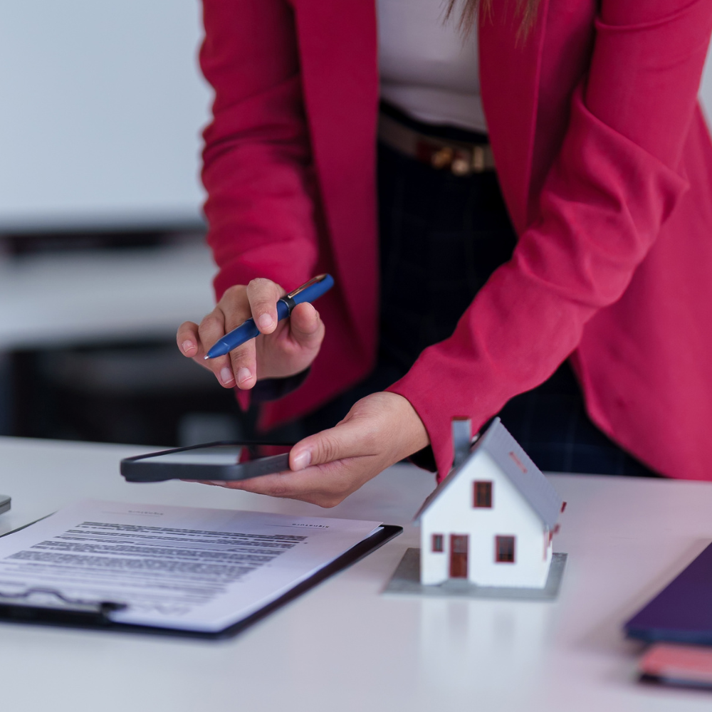 A person in a red blazer checks a smartphone next to a small model house and a contract on a white desk.