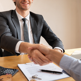 A person in a business suit shakes hands with another person over a contract on a wooden desk.