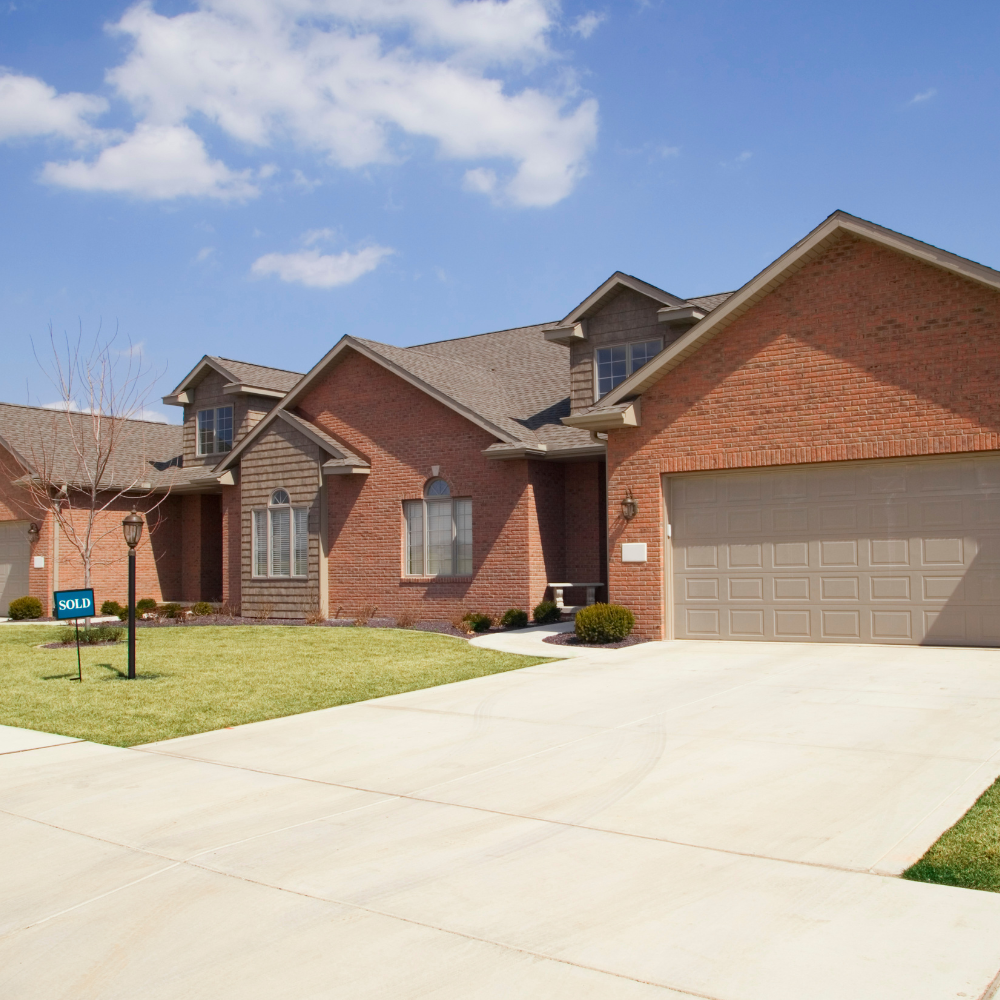 A brick suburban house with a two-car garage and a for-sale sign on the lawn under a blue sky.