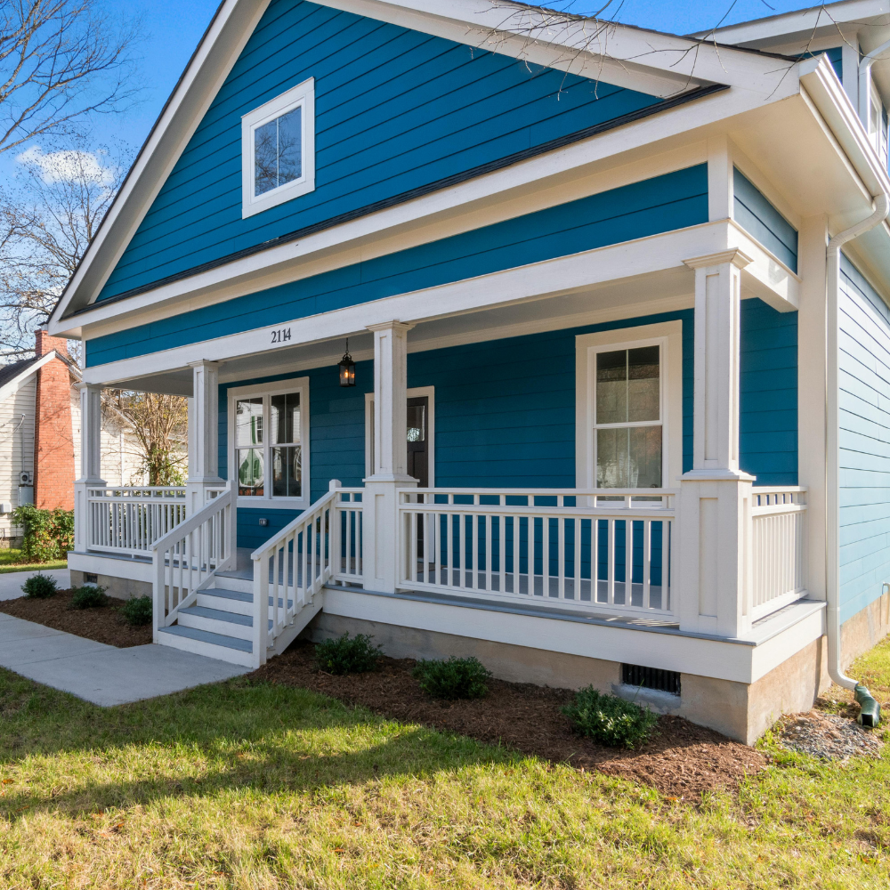 A teal, single-story house with a white wraparound porch, stairs, and a front yard under a clear blue sky.