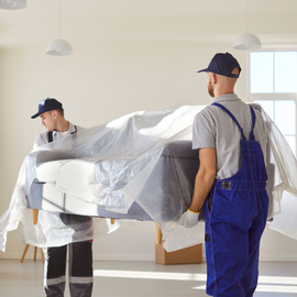Two movers in uniforms and caps carry a sofa wrapped in protective plastic inside an empty room.