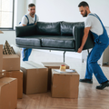 Two movers in blue overalls carry a black sofa through a room filled with cardboard moving boxes.