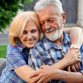 A couple embraces tenderly, smiling with their eyes closed, wearing casual plaid shirts in an outdoor setting.