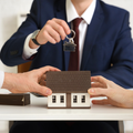 A person in a suit hands house keys over a miniature model house held by two other people on a table.