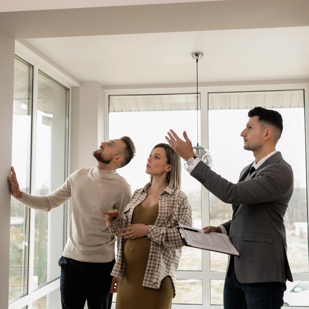 A real estate agent points out features of a bright room to a couple as they look toward the windows and ceiling.