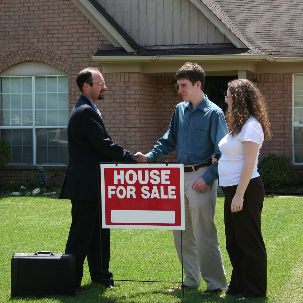A realtor in a suit shakes hands with a person in front of a house for sale sign on a lawn; another person stands nearby.