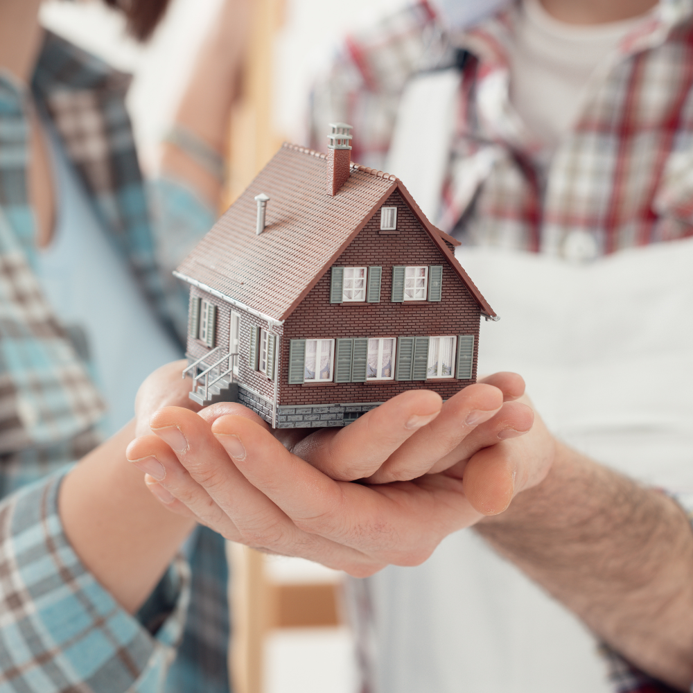 Two pairs of hands holding a small model of a brown house with a shingled roof, set against a blurred background.