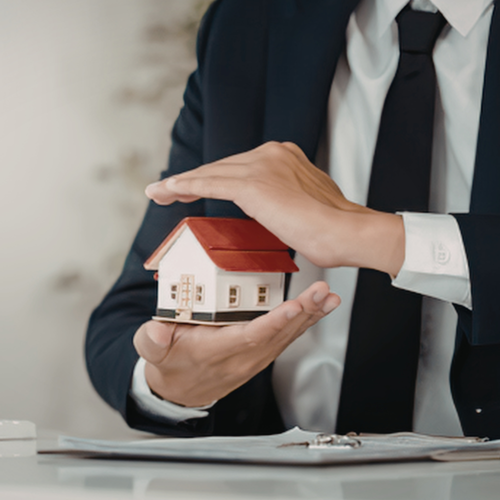 A person in a business suit protects a small model house with their hands, symbolizing real estate or insurance coverage.