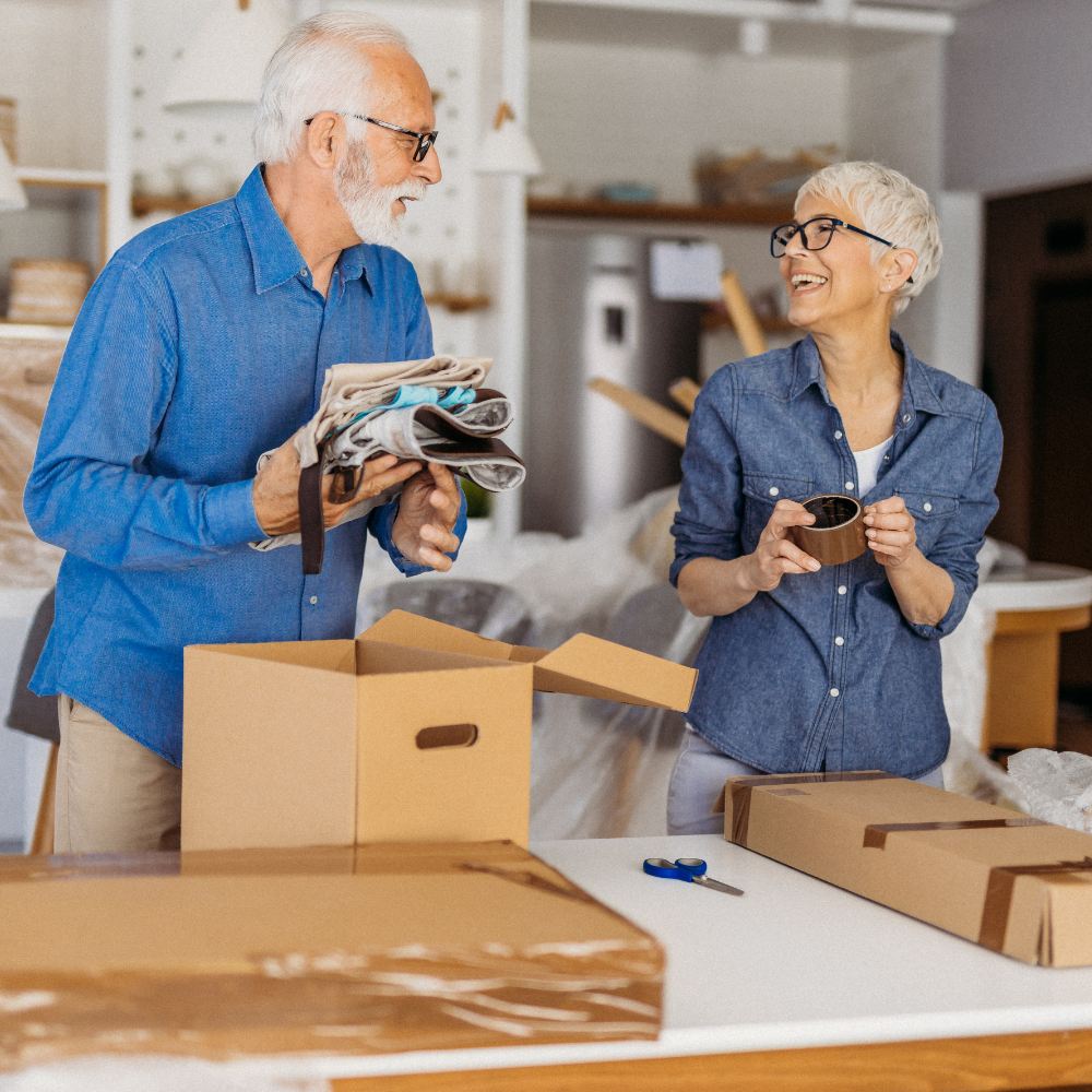 Two people in denim shirts packing items into cardboard boxes at a table in a home setting.
