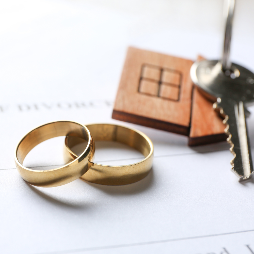 Two gold wedding rings rest on a legal document next to a key and a small wooden house icon.