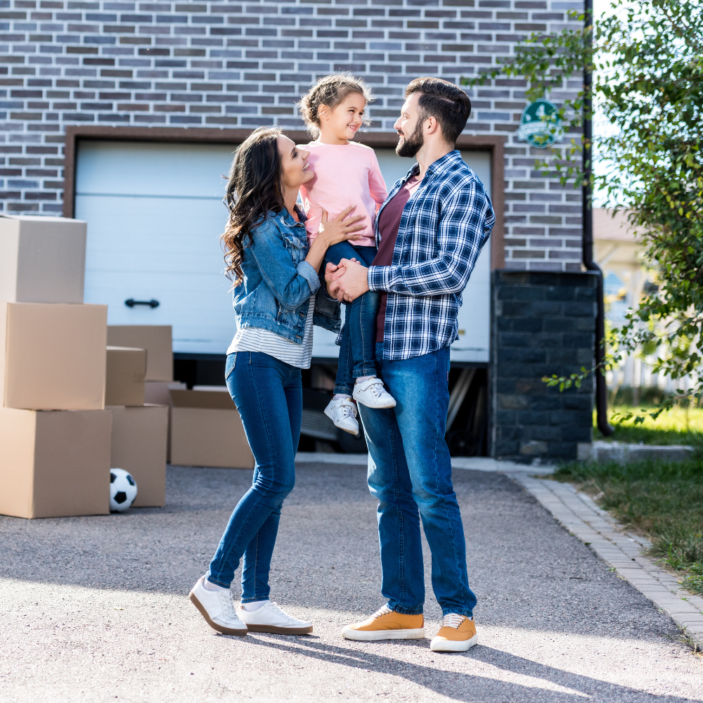A family stands in a driveway next to a garage with several cardboard moving boxes, smiling at one another.