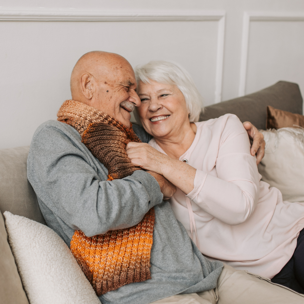 Two people sitting on a sofa, laughing and embracing while wrapped in a textured, multi-colored scarf.