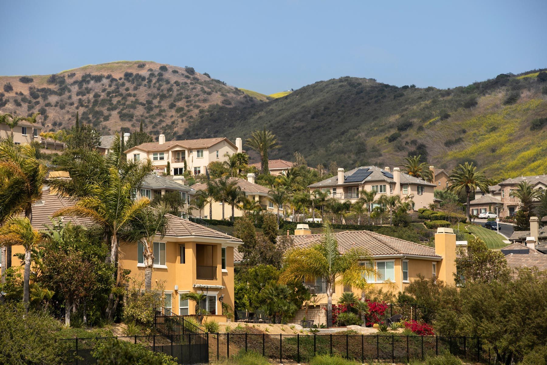 Stucco houses nestled on a hillside with dry, grassy slopes and scattered trees under a clear blue sky.