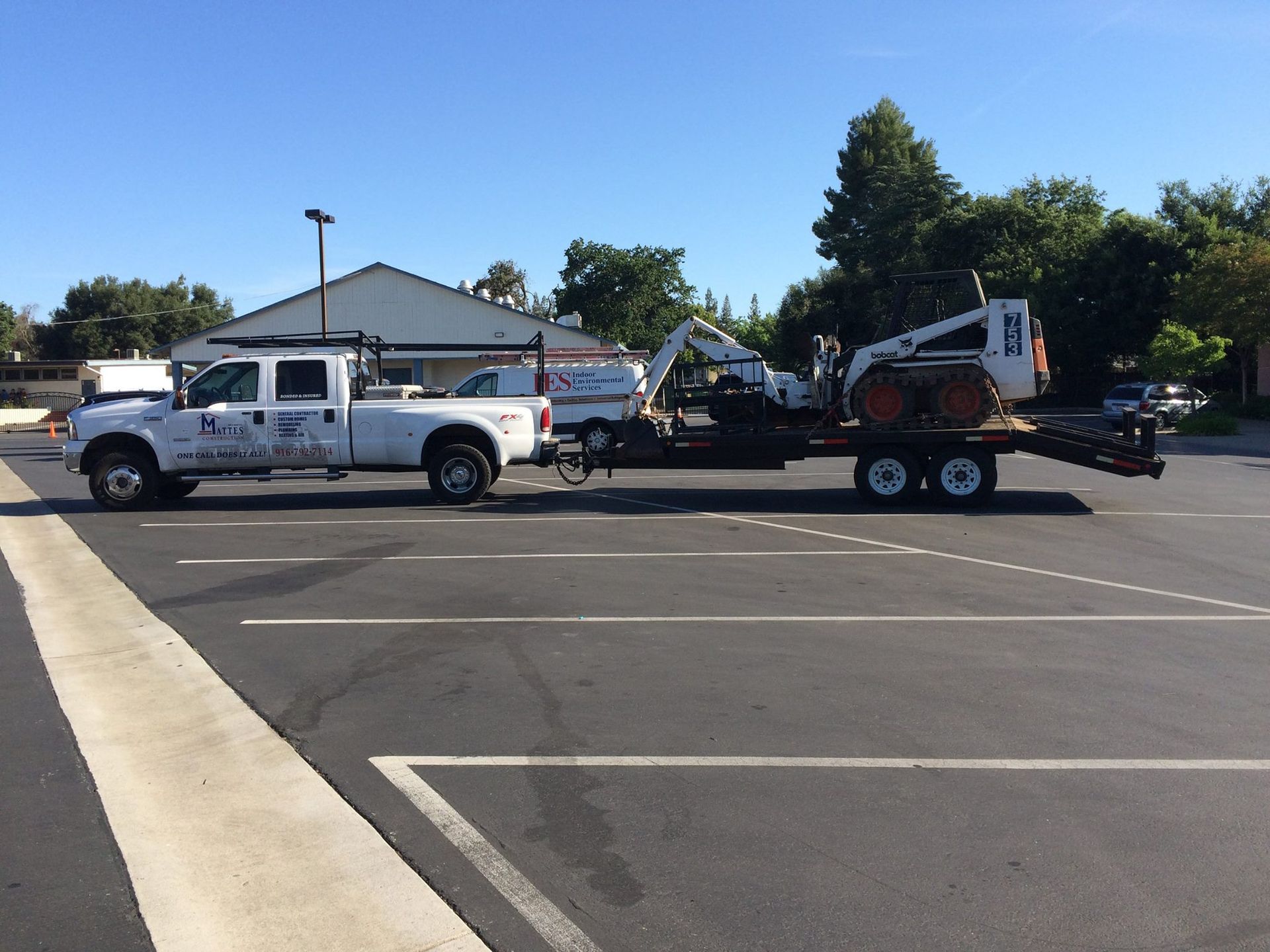 White truck towing a trailer with a Bobcat and excavator in a parking lot.