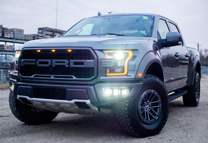Gray Ford Raptor truck with black accents, parked outdoors, lit headlights.