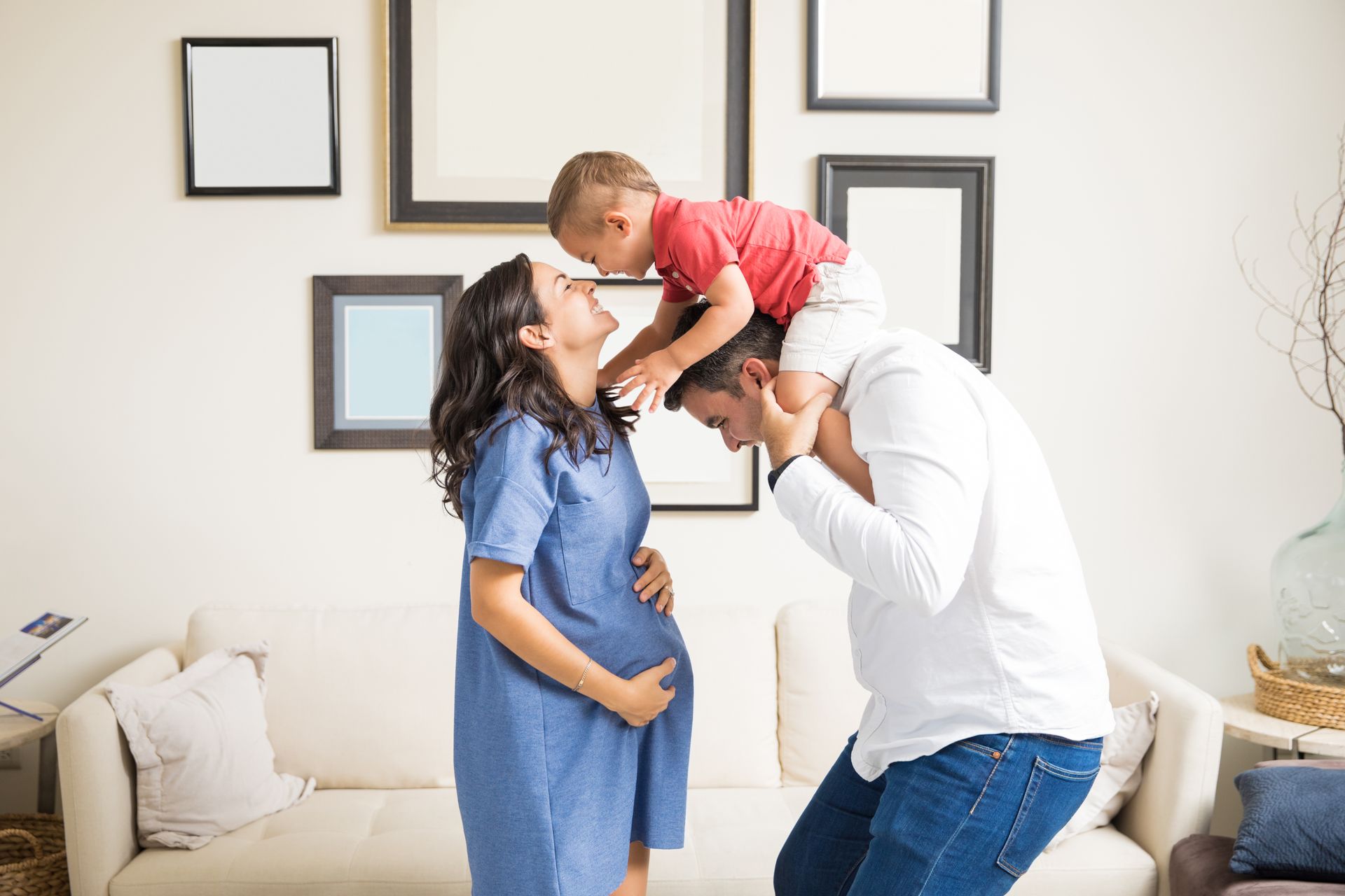 Pregnant person with partner, child on shoulders; family in living room with frames.