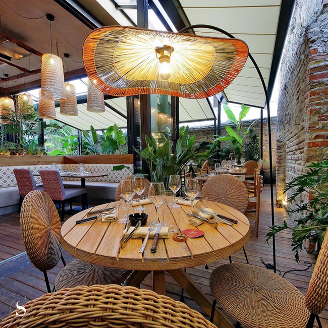 Rieten stoelen en ronde tafels op een lichte, met planten gevulde restaurantpatio.