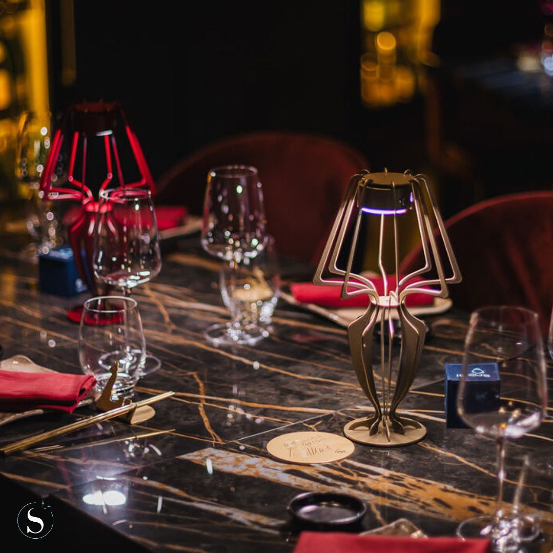 Een schemerig verlichte restauranttafel met wijnglazen en lampen met rode kappen op een marmeren blad.