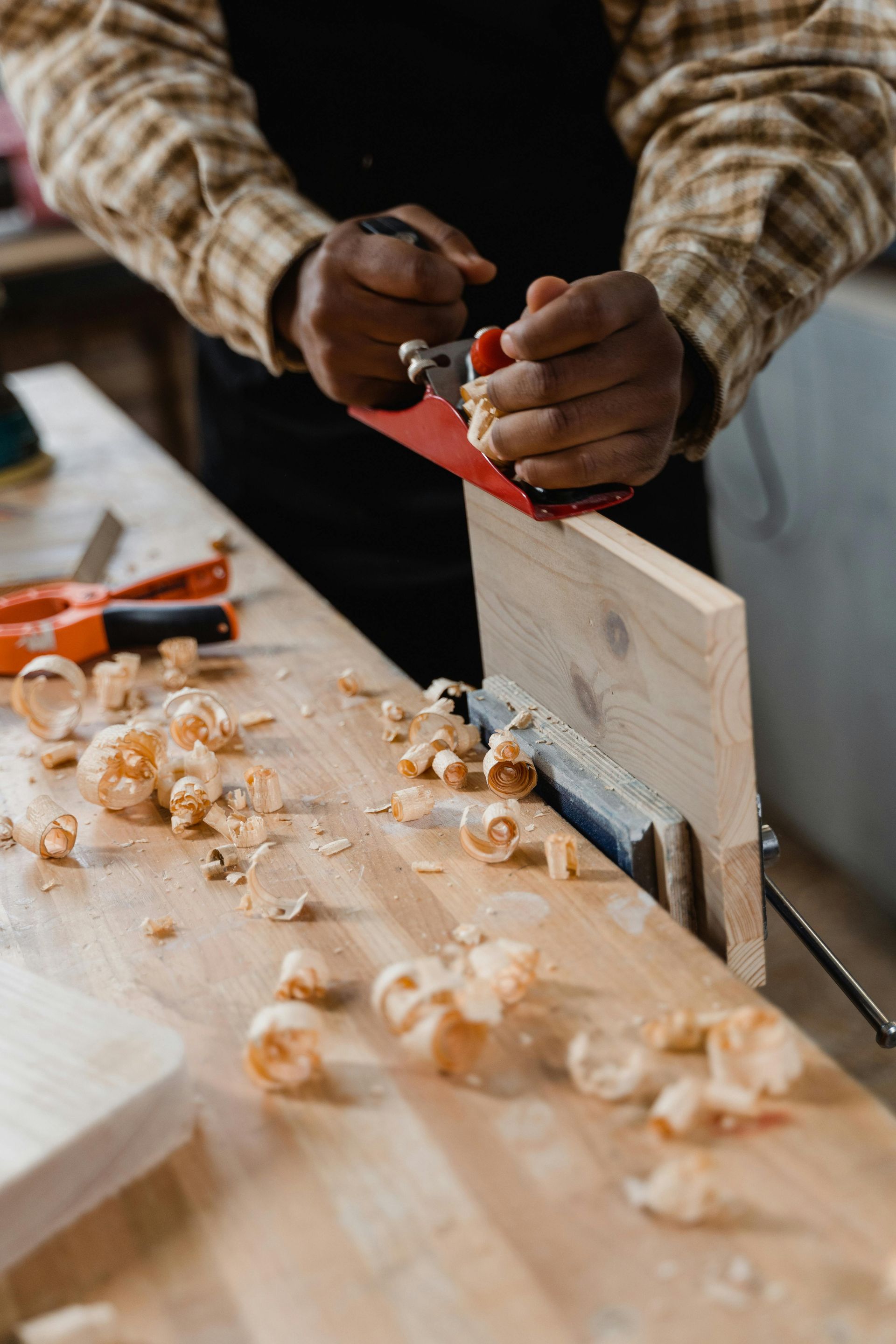 Hands using a red hand plane on a wooden board, with wood shavings scattered on the workbench.