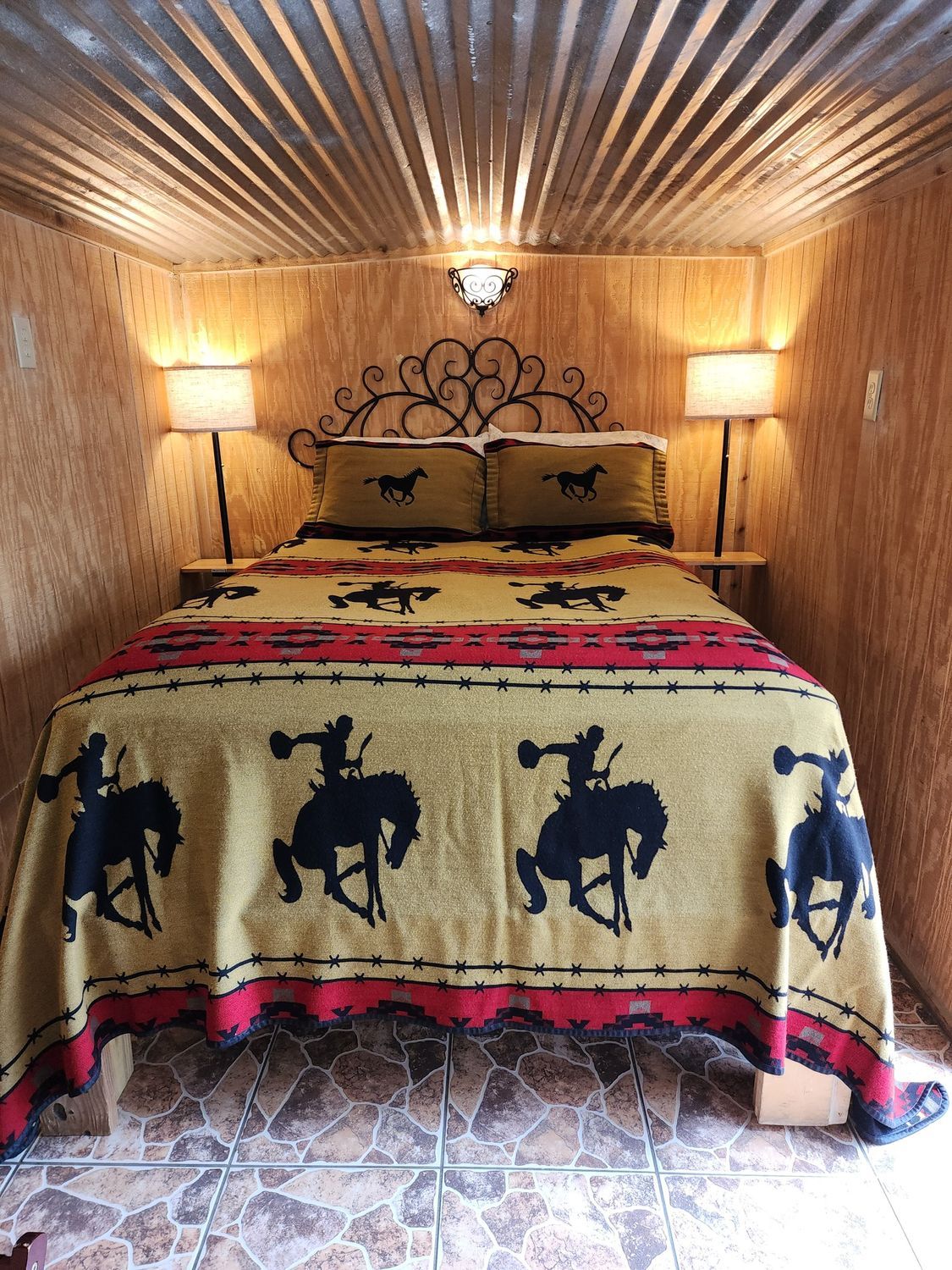 A bedroom with a bed , two lamps , and a blanket with horses on it.