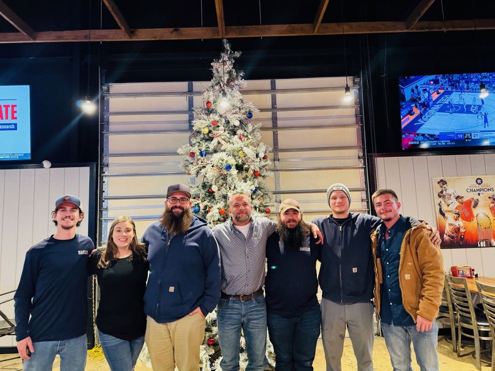 Group posing indoors in front of a decorated Christmas tree, with TVs and restaurant seating nearby.