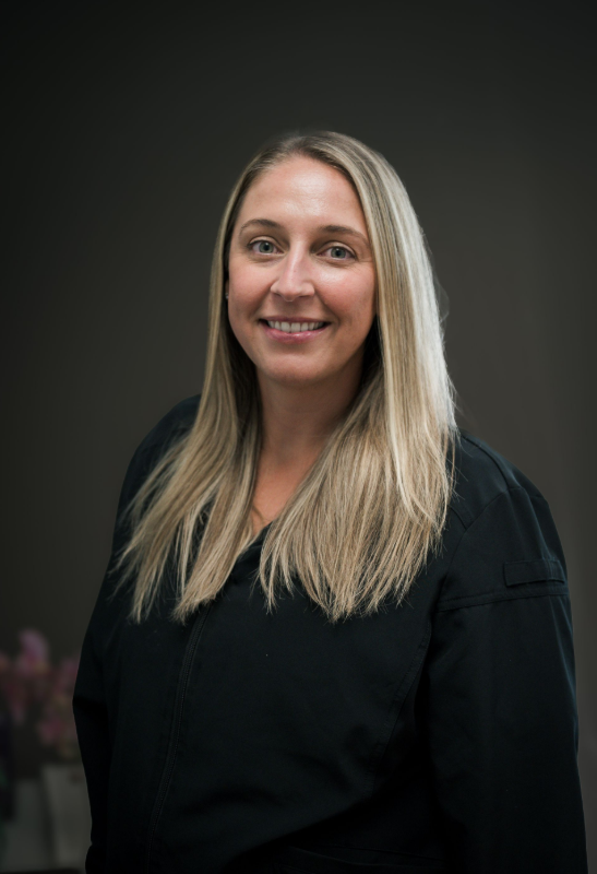 Woman with long blonde hair, wearing a black top, smiles in front of a neutral backdrop.