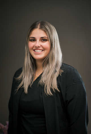 Woman with long, light-colored hair smiles at the camera, wearing a black jacket and top, with a gray background.