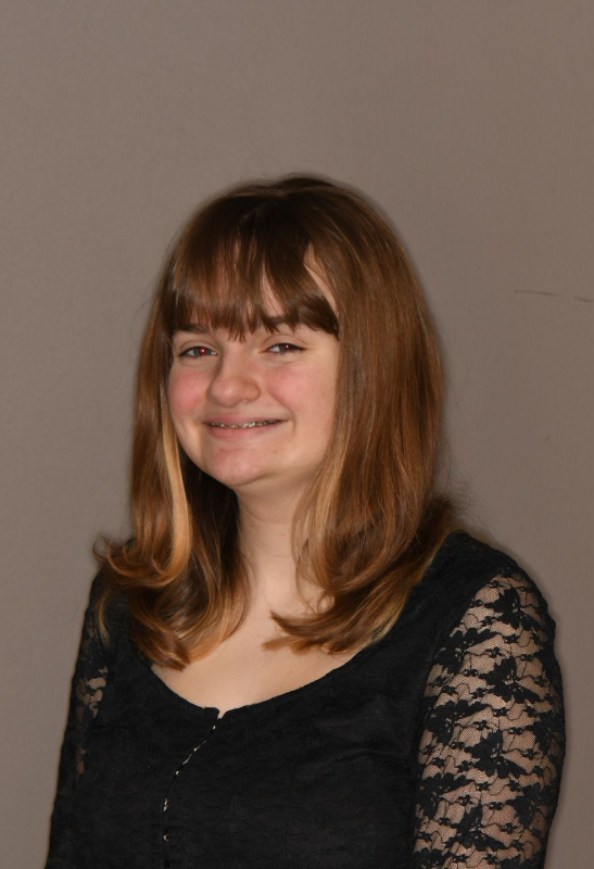 Woman with light brown hair and bangs smiles, wearing a black lace-sleeved top, and braces.