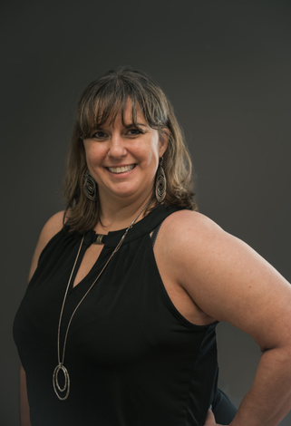 Woman in black sleeveless top smiles, hand on hip, against a gray background.