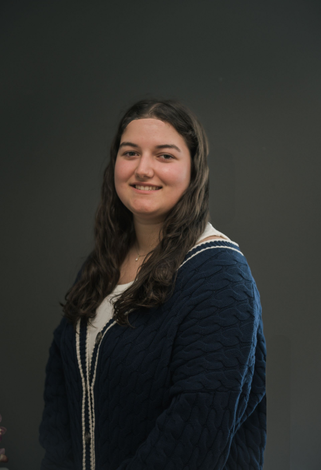 Woman with dark hair smiles, wearing a navy cardigan over a white top, against a gray background.