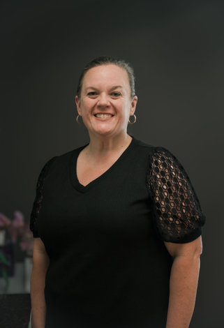 Woman smiling, wearing a black top with lace sleeves, standing in front of a dark gray wall.