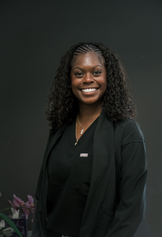 Smiling person in black medical scrubs and jacket, dark background.