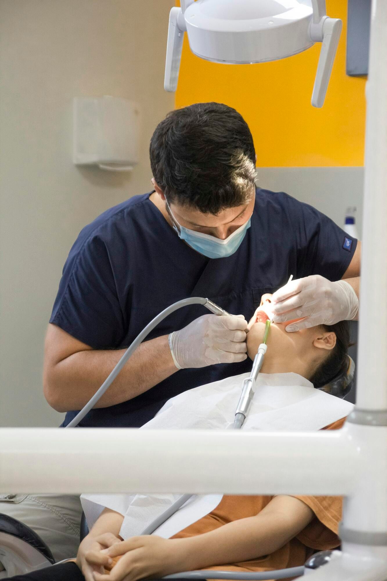 A dentist in a blue uniform and face mask performs a procedure on a patient in a dental chair under clinical lights.