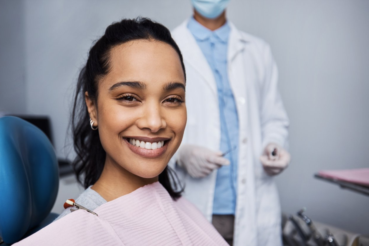 Woman smiling in a dental chair; dentist in the background wearing mask, gloves, and a lab coat.