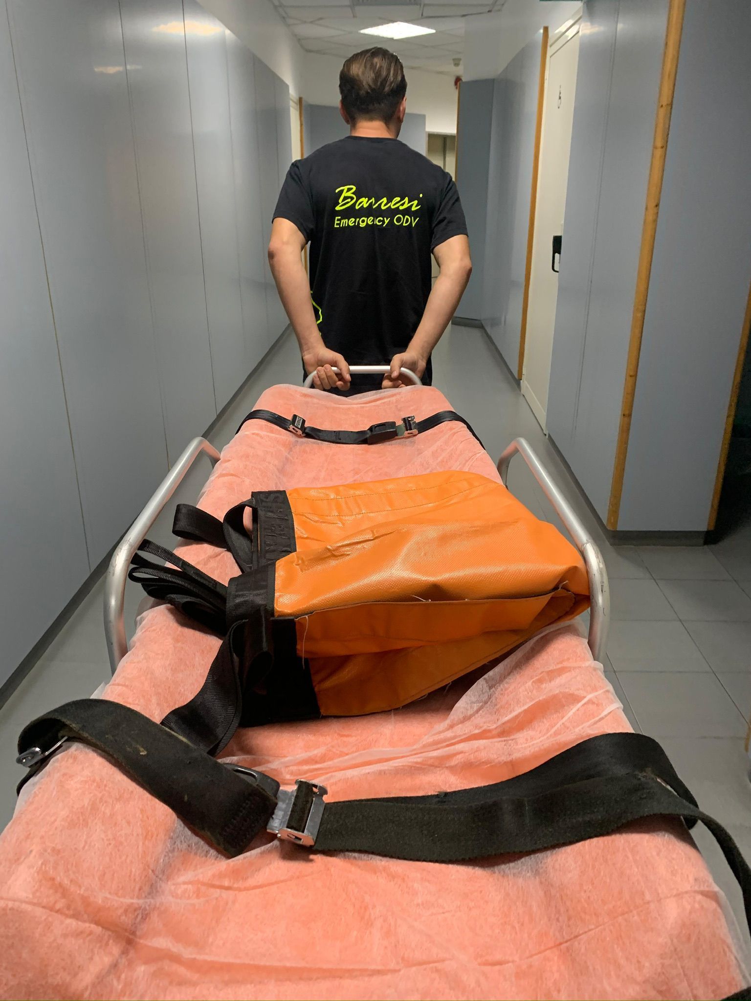 Persona che trascina una barella lungo un corridoio. Sulla barella è appoggiata una borsa.