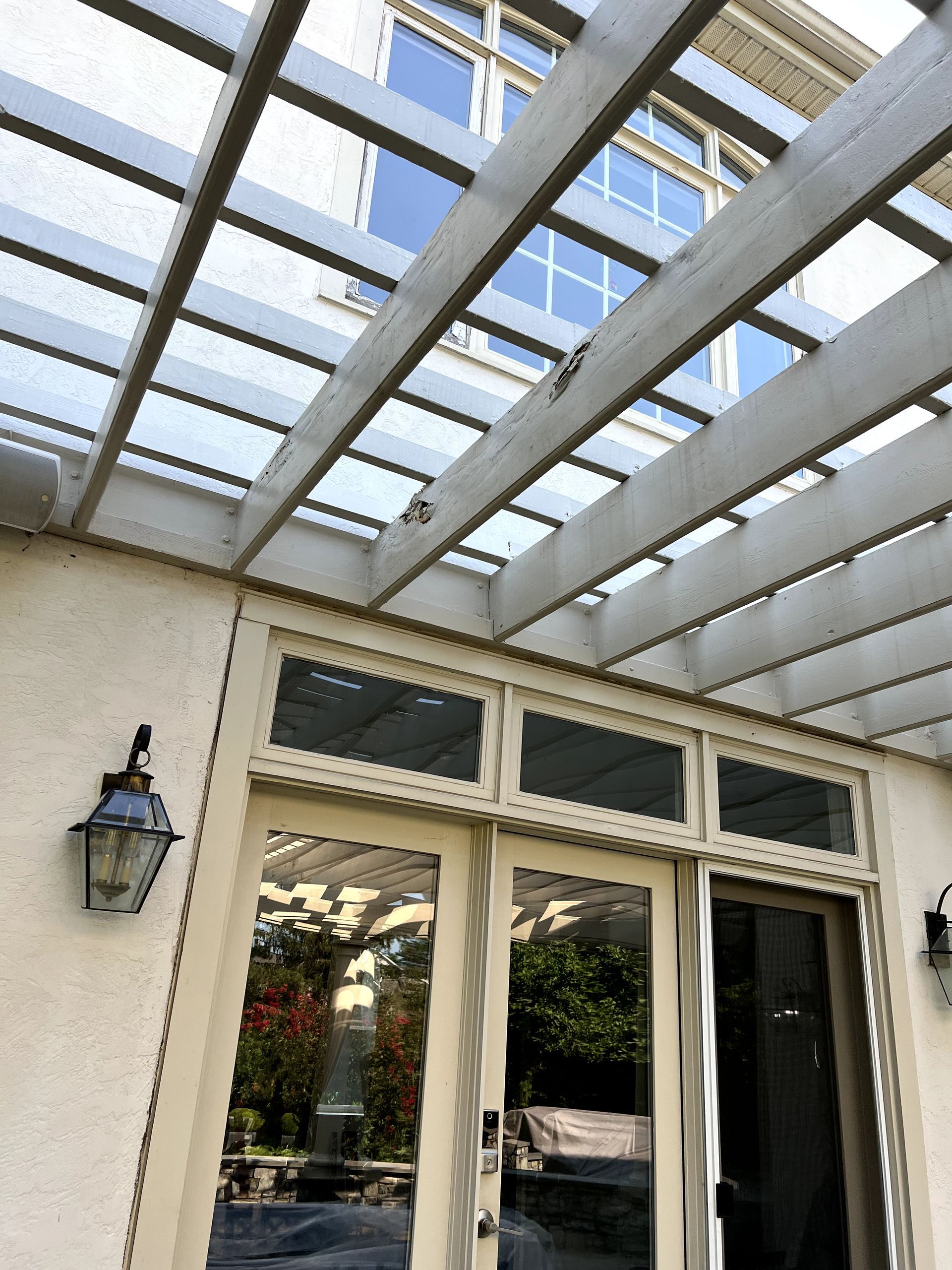 Exterior view of a pergola over sliding glass doors, with a lamp on the stucco wall.