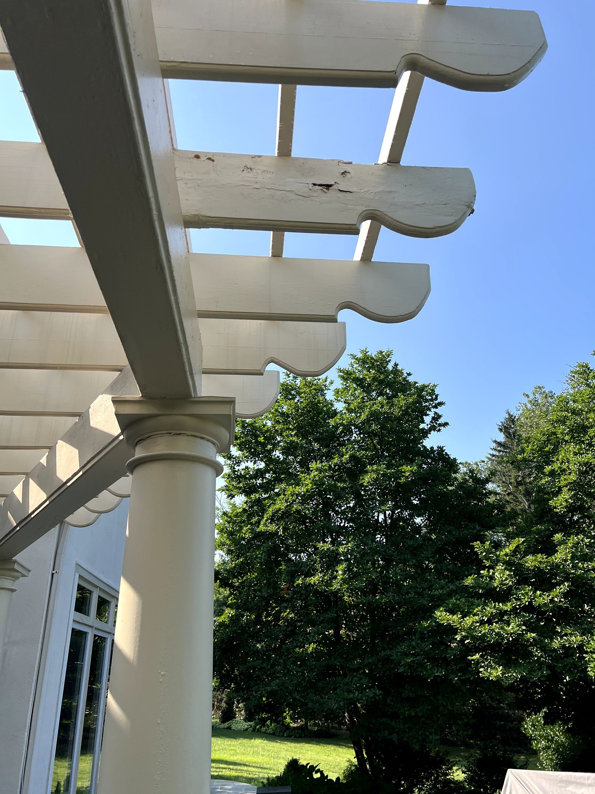 White pergola with decorative edges, supported by a column, trees and a clear blue sky.