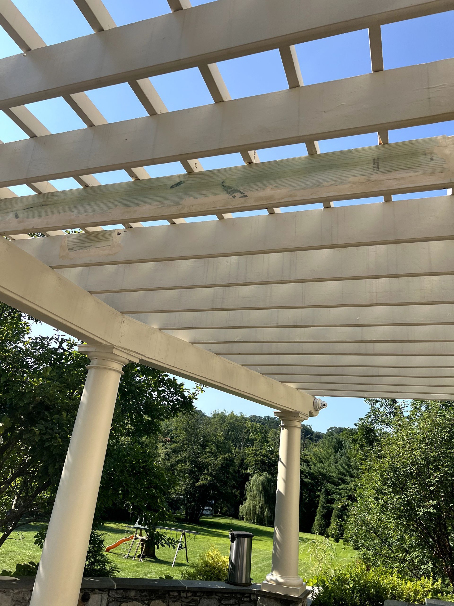 White pergola with visible green algae growth, overlooking a green yard and trees.