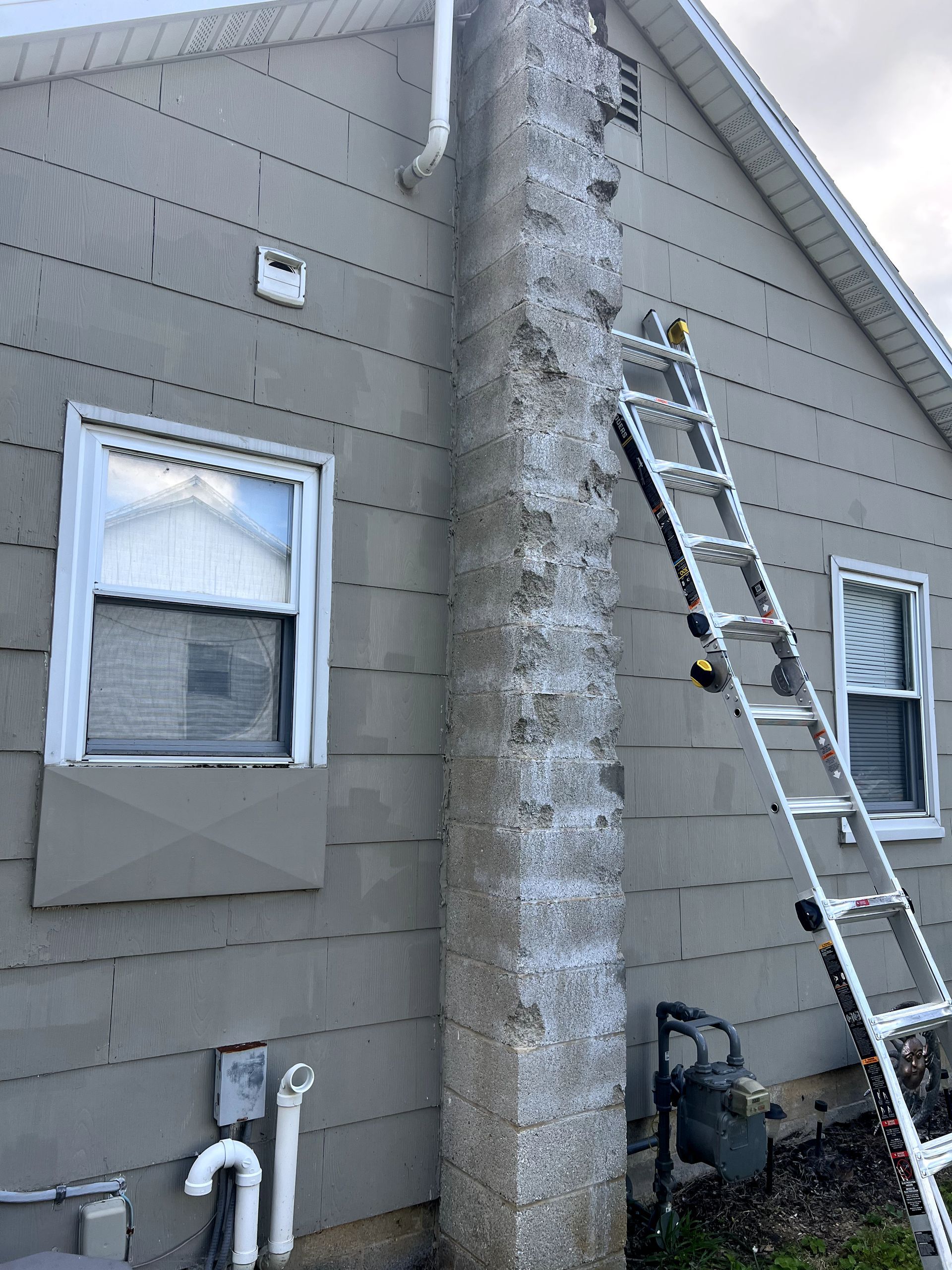Ladder against a weathered chimney on a gray-sided house, two windows visible.