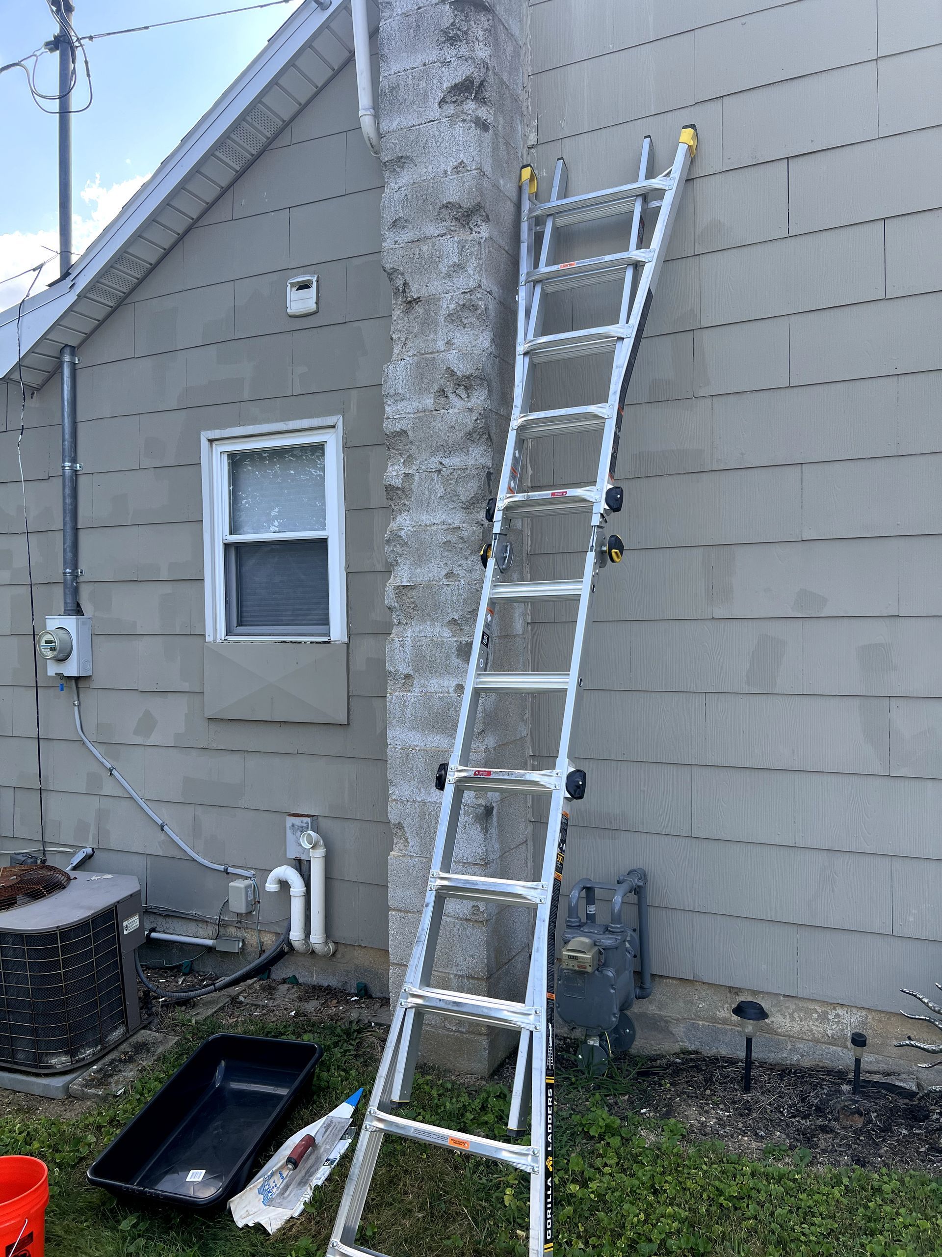 A ladder leans against a house's textured wall next to a chimney, with tools and a tray at the base.