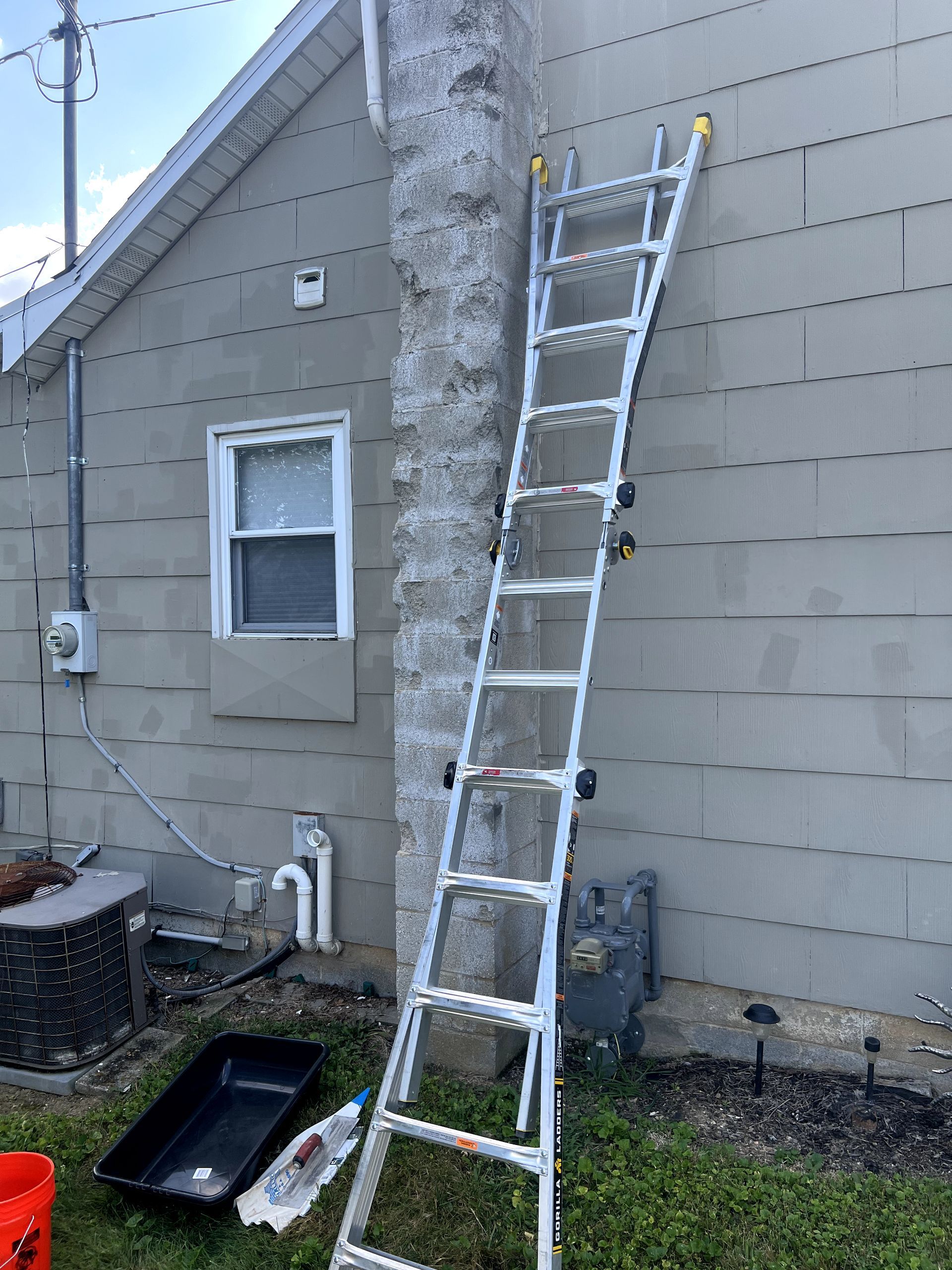 Ladder leaning against a house wall next to a chimney, with tools on the ground.