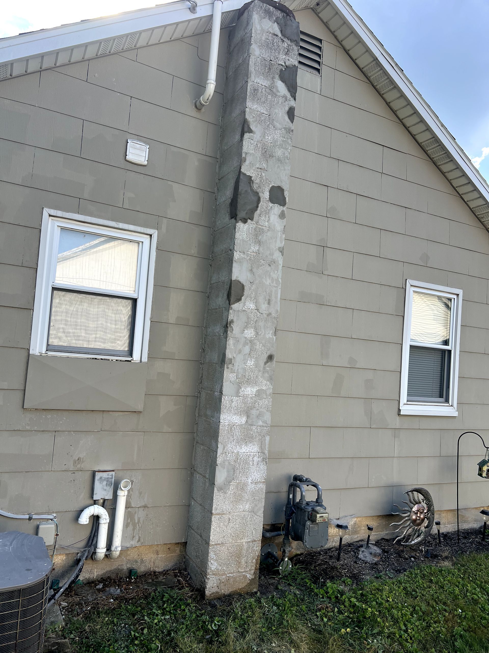 Chimney leaning away from a house with gray siding and two windows.