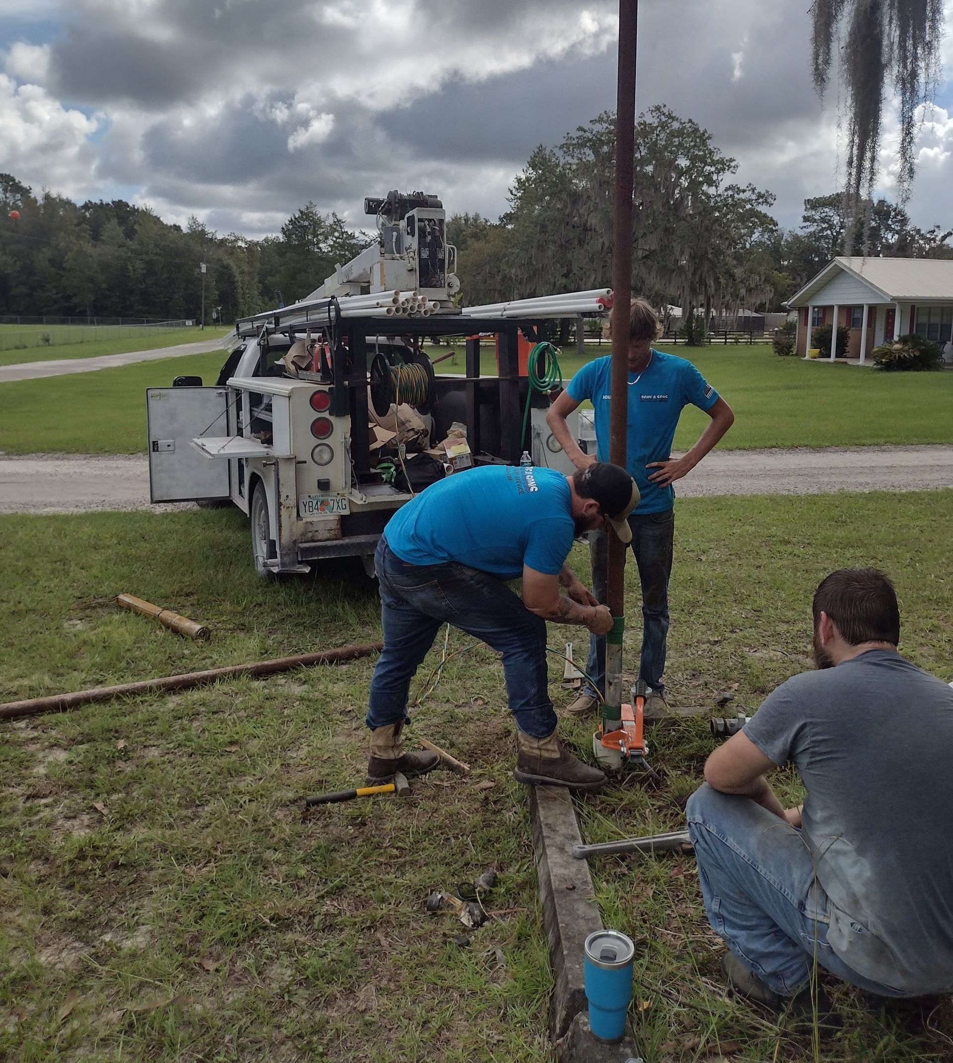 Three people working on a pole in a grassy yard, with a truck and house in the background.
