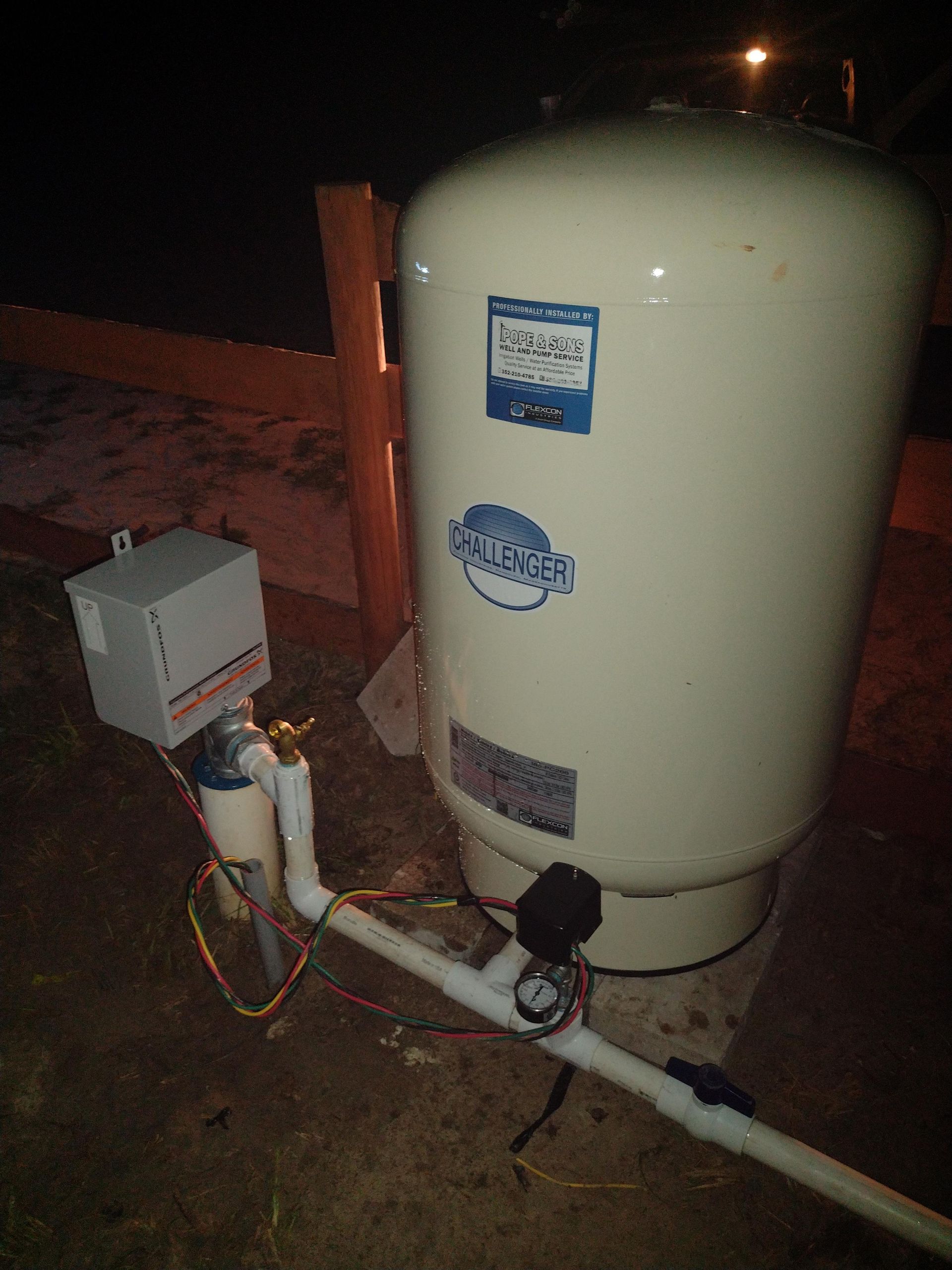 Well water system with pressure tank, pipes, gauges, and electrical box outdoors at night.