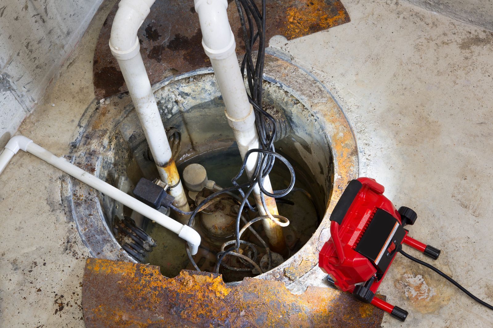Sump pit with pipes, pump, and cords, lit by a red work light. Brown rusty metal surrounds.