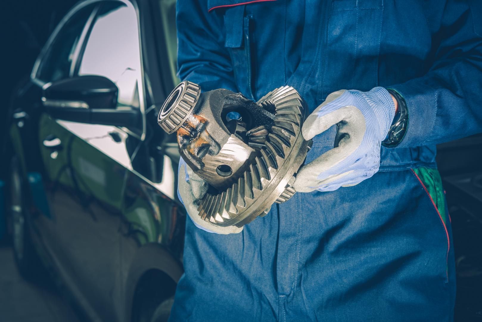 Mécanicien en salopette bleue tenant un différentiel de voiture près d'une voiture noire.