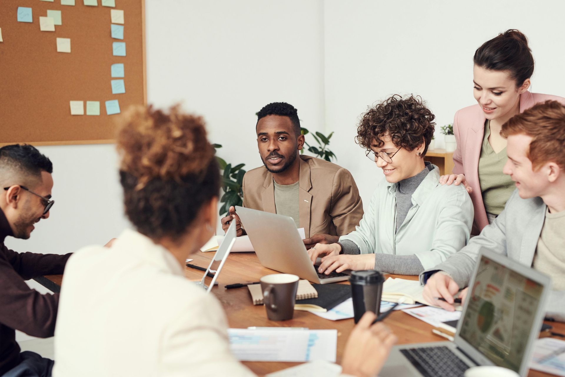 People at a table looking at laptops. Some are smiling, working in an office with a corkboard.