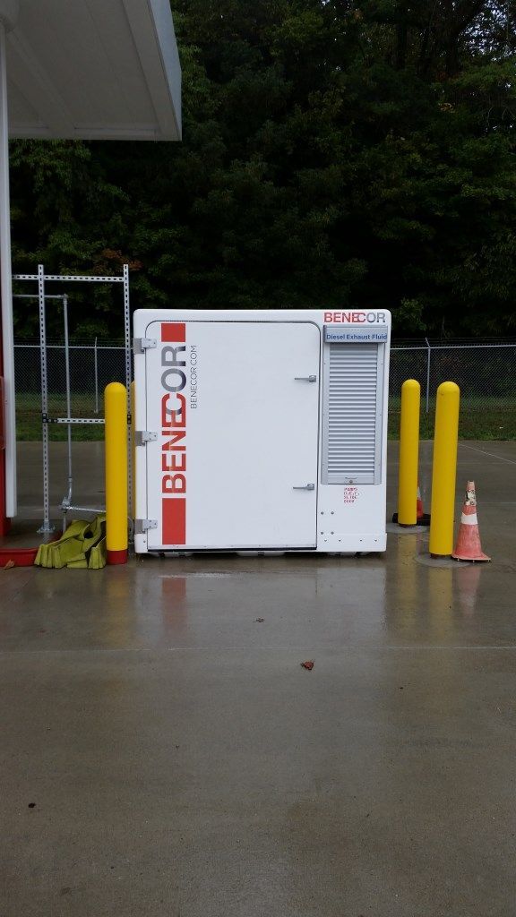 White BENECOR generator box, red and black logo, yellow bollards, wet concrete at gas station.