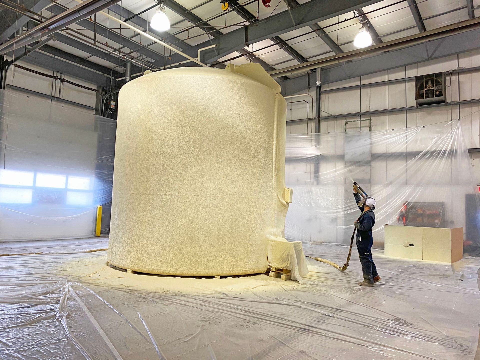 A person spraying foam on a large, cylindrical object inside a warehouse.