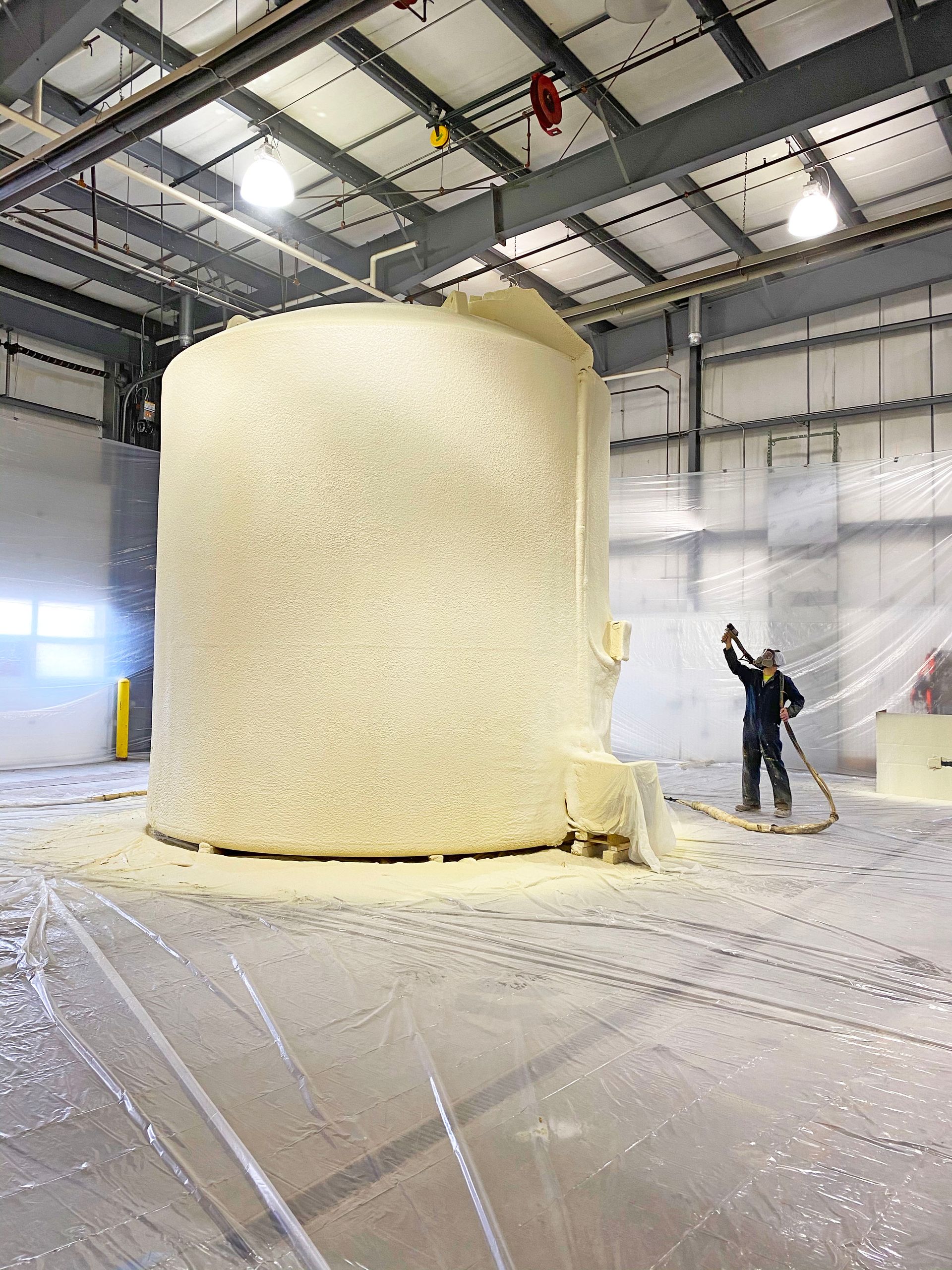 Man sprays foam onto a cylindrical structure in a warehouse. Foam covers the structure and floor.